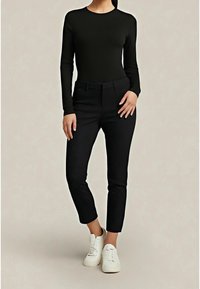 Femme portant un haut noir moulant à manches longues, un pantalon noir court et des baskets blanches, debout devant un fond neutre.