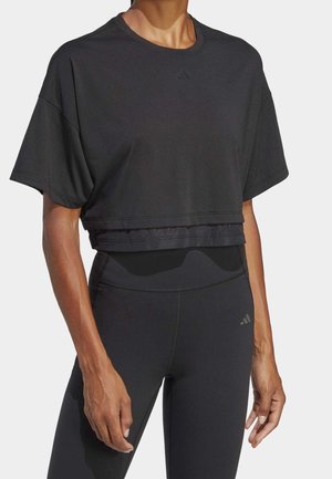 Kobieta ubrana w czarny krótki top z krótkim rękawem i wysokowciskane czarne legginsy, stojąca z ramionami swobodnie zwisającymi wzdłuż ciała.