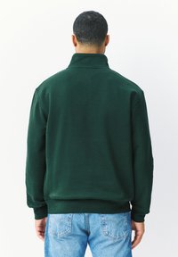 Zelená mikina s vysokým límcem, vyrobená z měkké látky. Má žebrované manžety a spodní lem. Model nosí světle modré džíny.