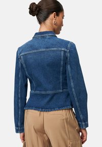 Donkerblauwe denimjas met een getailleerd ontwerp, spitse kraag en zichtbare stiksels. De jas heeft lange mouwen en een gebogen zoom.
