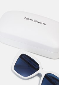 Lunettes de soleil Calvin Klein Jeans avec des montures en plastique blanc, des verres bleus et un étui rigide blanc assorti portant le logo de la marque.