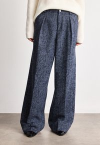 Calças largas em tecido texturizado azul escuro com pregas, apresentando um fecho frontal e um ajuste personalizado, combinadas com um suéter de cor clara.