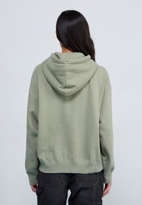 Hooded sweatshirt in soft olive green fabric, featuring a relaxed fit, ribbed cuffs, and a drawstring hood. Detailed stitching along seams.