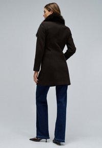 Abrigo de lana marrón con cuello de piel, diseño ajustado, mangas largas, longitud a mitad del muslo, combinado con jeans de mezclilla oscuros de corte acampanado y zapatos de punta.
