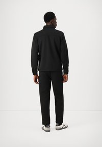 Jacket preta com fecho éclair e gola alta, combinada com calças pretas a condizer e ténis brancos. Textura suave, design minimalista.