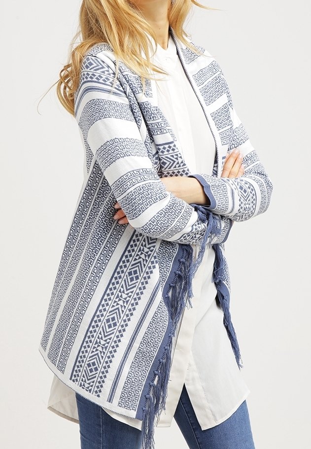 Femme portant un cardigan ouvert bleu et blanc à motifs avec des franges sur une chemise blanche et un jean bleu, bras croisés.