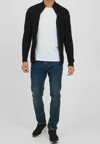 Czarny prążkowany kardigan na zamek błyskawiczny, noszony na białej koszulce, w zestawie z niebieskimi dżinsami i czarnymi butami sportowymi. Luźne dopasowanie.