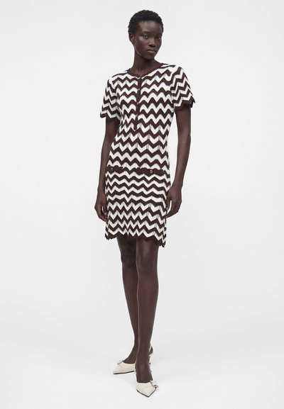 Femme portant une robe courte à manches courtes, longueur genou, avec un motif en zigzag marron et blanc et des talons blancs pointus avec des nœuds.