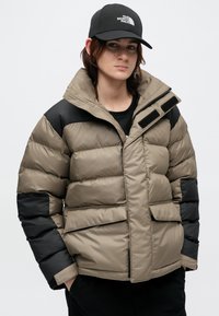 Kurtka puchowa w beżowym kolorze z czarnymi akcentami, wyposażona w wysoki kołnierz, zamknięcie na zamek oraz dwie przednie kieszenie. Wykonana z pikowanego, gładkiego materiału.