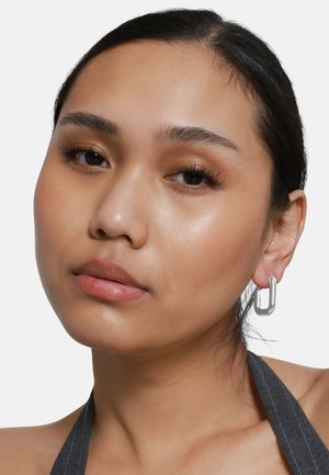 Orecchini in metallo argentato a forma di U con una finitura liscia, indossati da un modello con trucco neutro e capelli scuri. Design minimale e texture elegante.