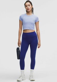 Nő világoslila cropped topban, sötétkék leggingsben, fehér zokniban és sportcipőben, fekete vízpalackot tartva, sima háttér előtt áll.