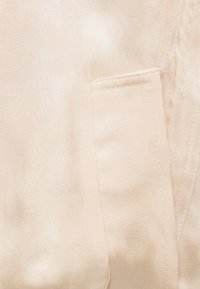 Κοντινό πλάνο μπεζ υφάσματος που δείχνει μια μικρή ραμμένη τσέπη με ορατές ραφές και υφή σε λείο φόντο.