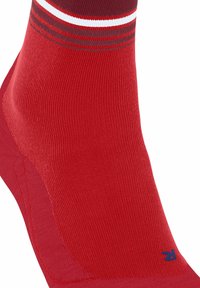 Primo piano di un calzino rosso alla caviglia con tallone e punta rinforzati, caratterizzato da strisce bianche e rosso scuro vicino al bordo superiore e una piccola "R" blu sulla punta.