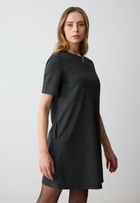 Vrouw met licht haar in een eenvoudige donkergrauwe jurk met korte mouwen, staande tegen een effen lichte achtergrond.