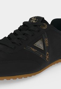Baskets noires Guess avec lacets texturés, plaque logo dorée, détail latéral à motifs et semelle en caoutchouc marron.