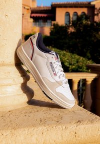 Baskets Reebok blanches et beiges avec une doublure intérieure noire reposant contre un pilier en pierre sur une terrasse extérieure ensoleillée.
