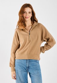 Beige cipzáras pulcsi magas gallérral, laza fazonnal, bordázott mandzsettákkal és texturált anyagból, kék farmernadrággal párosítva.