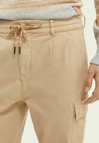 Beige bukser med knap og snoreliv, cargo-lomme på låret og hånden hvilende nær lommen, parret med en flerfarvet strikketrøje.