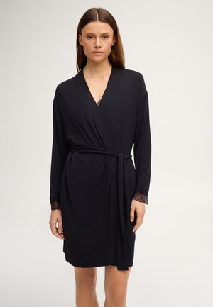 Vrouw draagt een zwarte wikkeljurk tot op de knie met lange mouwen en met kant afgewerkte manchetten, gestrikt in de taille met een bijpassende riem.