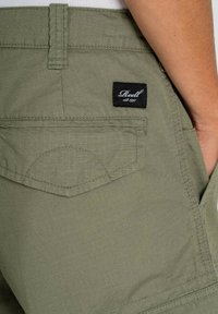 Pantalones cortos verde oliva con textura a cuadros, que presentan un bolsillo trasero y una etiqueta negra con texto blanco. La tela es ligera.