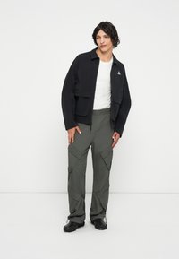 Jaqueta preta curta com dois bolsos da frente, combinada com calças cargo largas em tom verde-oliva com fechos de zíper e múltiplos bolsos. Calçado preto.