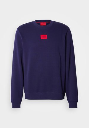 Navy blue sweatshirt with round neckline, long sleeves, ribbed cuffs, and a red rectangular logo patch on the front. Soft fabric texture.
