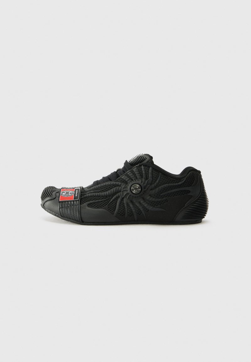 Chaussure de sport noire avec un design texturé, un motif de soleil en relief, un embout en caoutchouc et un écusson de logo bien visible sur le côté.