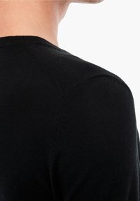 Gros plan sur l'épaule et le cou d'une personne portant un pull noir à col rond sur fond blanc.