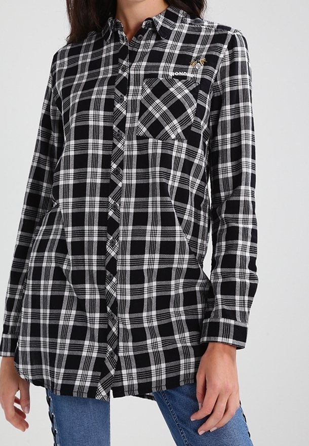 Chemise à manches longues à carreaux noir et blanc avec poche avant et fermeture à boutons, portée sur un jean bleu.