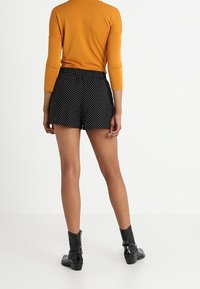 Černé šortky s puntíky a uvolněným střihem, vyrobené z lehkého materiálu. V kombinaci s přiléhavým oranžovým tričkem s dlouhým rukávem a černými kotníkovými botami.