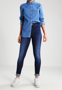 Camisa de mezclilla de botones en azul claro, con un bolsillo en el pecho; combinada con jeans skinny de índigo oscuro y zapatillas blancas, con los puños enrollados.
