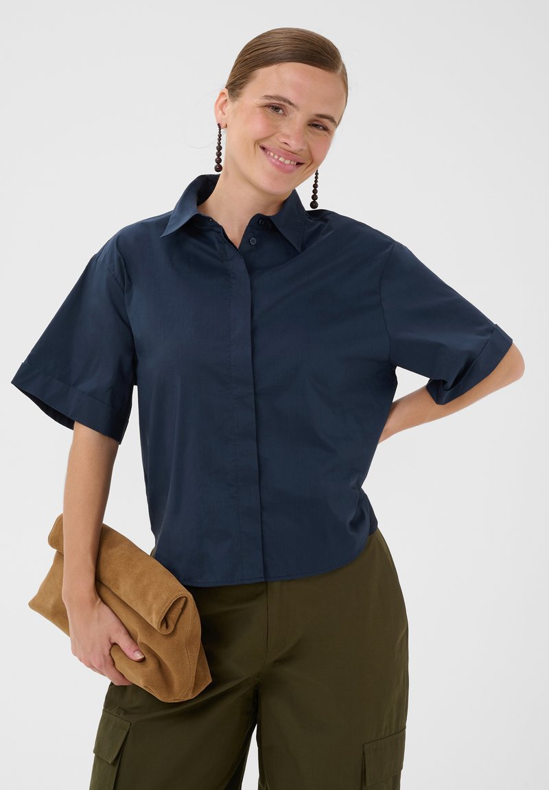 Femme souriante vêtue d'une chemise bleu marine à manches courtes, pantalon cargo kaki, tenant une pochette beige pliée, portant de longues boucles d'oreilles perlées, sur un fond uni.
