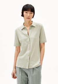 Camicia short-sleeve verde chiaro con bottoni sul davanti e colletto a punta, realizzata in tessuto liscio, indossata con pantaloni grigi a righe.