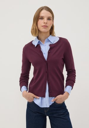 Frau mit kurzen blonden Haaren, die eine violette, geknöpfte Strickjacke über einem blau gestreiften Hemd und dunklen Jeans trägt und mit den Händen in den Taschen steht.