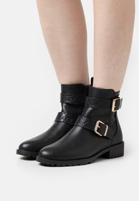 Botas negras hasta el tobillo con una correa texturizada, hebillas y una suela plana. Presentan un acabado de cuero liso y una suela exterior resistente y con tacos.