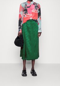 Blusa colorida con patrones abstractos; falda de satén verde con estampado de logo; bolso negro esponjoso; mocasines negros.