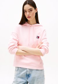 Sweatshirt à capuche rose clair avec un écusson logo �à l'avant, des poignets côtelés et une poche kangourou. Associé à un jean délavé bleu clair.