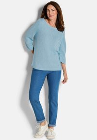 Hellblaue Strickpullover mit 3/4-Ärmeln und rundem Ausschnitt, kombiniert mit hochgerollten blauen Jeans und weißen Sneakers. Die Textur weist ein dezentes Muster auf.