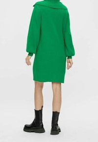 Robe en tricot verte avec un large col et de longues manches ballon, associée à des bottes noires à semelles épaisses, vue de dos.