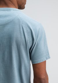 Man in een lichtblauw, kortärmig overhemd, gezien van achteren tegen een effen lichtgrijze achtergrond.