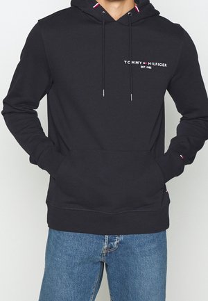 Mand iført sort Tommy Hilfiger-hoodie med frontlomme og blå jeans, hænderne i lommerne, viser logoet og mærkets detaljer på brystet og ærmerne.