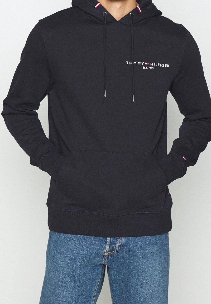 Homme portant un sweat à capuche noir Tommy Hilfiger avec poche avant et jean bleu, mains dans les poches, montrant le logo et les détails de la marque sur la poitrine et les poignets.