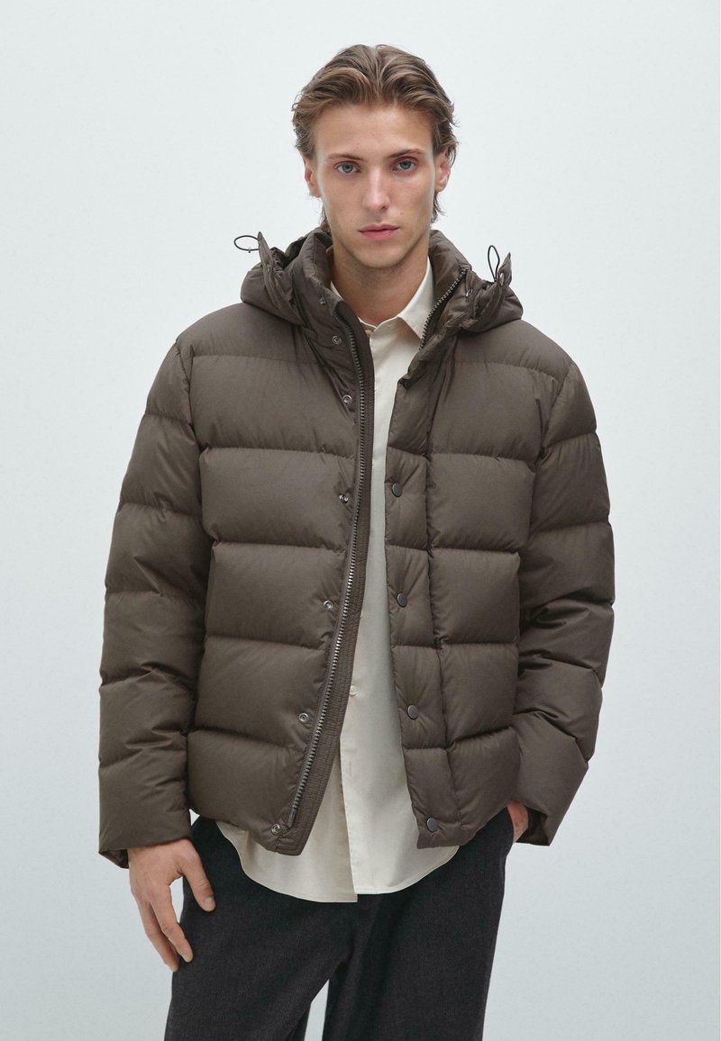 Hnědá prošívaná péřová bunda se zapínáním na zip, patenty a kapucí. Má velké prošívané části pro izolaci a hladký povrch.