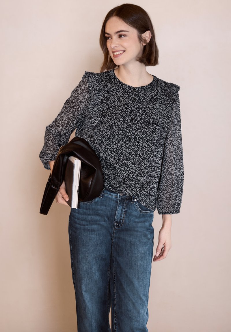 Jeune femme souriante, portant une blouse à motifs noirs et blancs avec un jean bleu, tenant une veste noire et un carnet sur un fond uni.