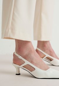 Talons en cuir blanc avec un bout pointu, un petit talon aiguille et un détail découpé sur le côté, positionnés contre un pantalon de couleur claire.