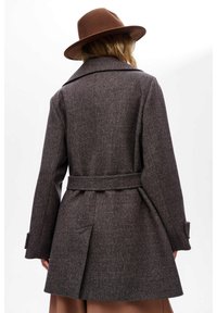 Bruine visgraatjas met een ceintuur in de taille, brede revers en knoopmanchetten, gecombineerd met een bruine vilten hoed.