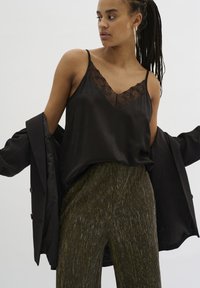 Camisola nera in satin con pizzo, abbinata a pantaloni larghi verde oliva scintillanti. La modella indossa un blazer nero.