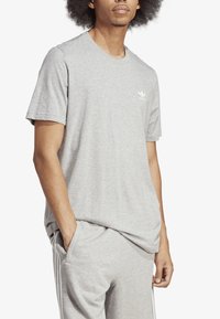 Šedé bavlněné tričko s kulatým výstřihem, krátkými rukávy a bílým logem na levé straně hrudi. Tkanina má hladkou, jemnou strukturu.