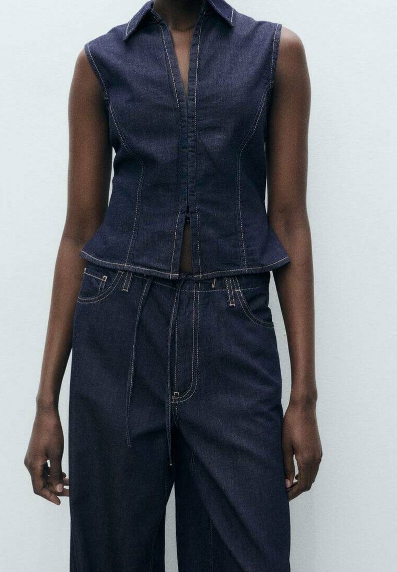 Haut sans manches en denim bleu foncé avec un corsage ajusté, un col et des coutures décoratives. Associé à un pantalon en denim taille haute à jambes larges.