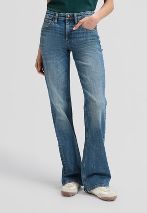 Person trägt blaue Schlagjeans, weiße Sneakers und ein dunkelgrünes Oberteil, eine Hand in der Tasche, steht vor einem einfachen hellen Hintergrund.